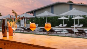 A wooden table with two glasses of amber-colored drinks, a bottle, and a vase with flowers. In the background, there are lounge chairs and umbrellas near a pool area. - V Luxury Suites (Corfu)