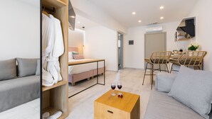 A modern hotel room with a sofa, a wooden coffee table with wine glasses, a wardrobe, a bed, and a dining area with chairs. - V Luxury Suites (Corfu)