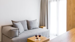 A modern hotel room with a grey sofa, a wooden side table with two glasses of red wine, and a large window with sheer curtains. - V Luxury Suites (Corfu)