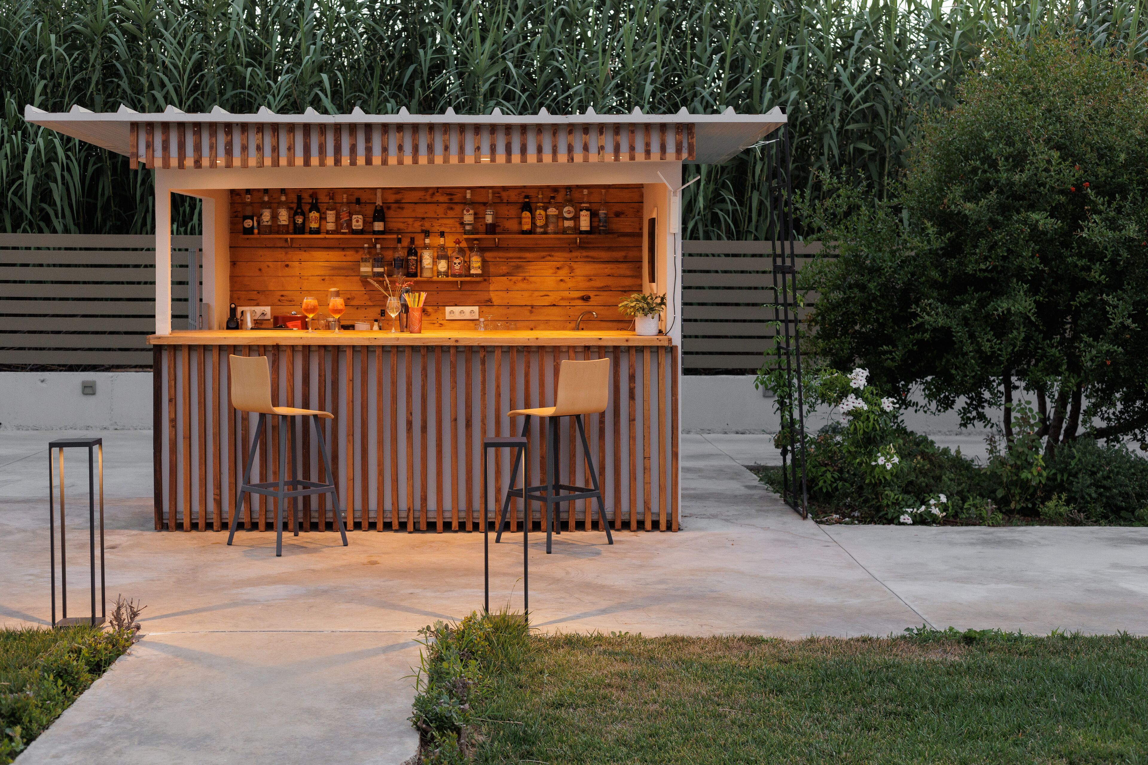 Een houten bar met twee krukken, een plank vol flessen en een kleine plant.