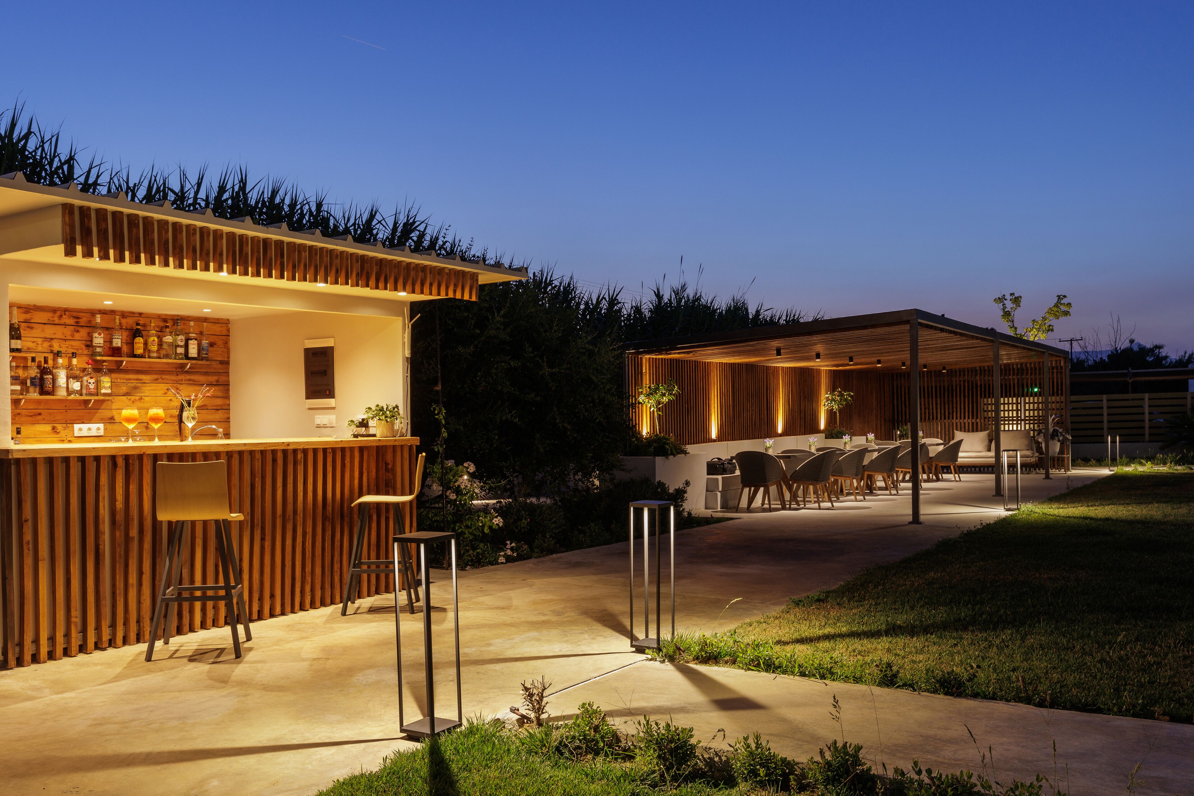 Un área de bar moderna al aire libre con paneles de madera, taburetes de bar y una zona de estar con mesas y sillas.