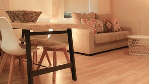 Een moderne woonkamer met een houten eettafel, witte stoelen, een bank met gekleurde kussens en een hangplant.