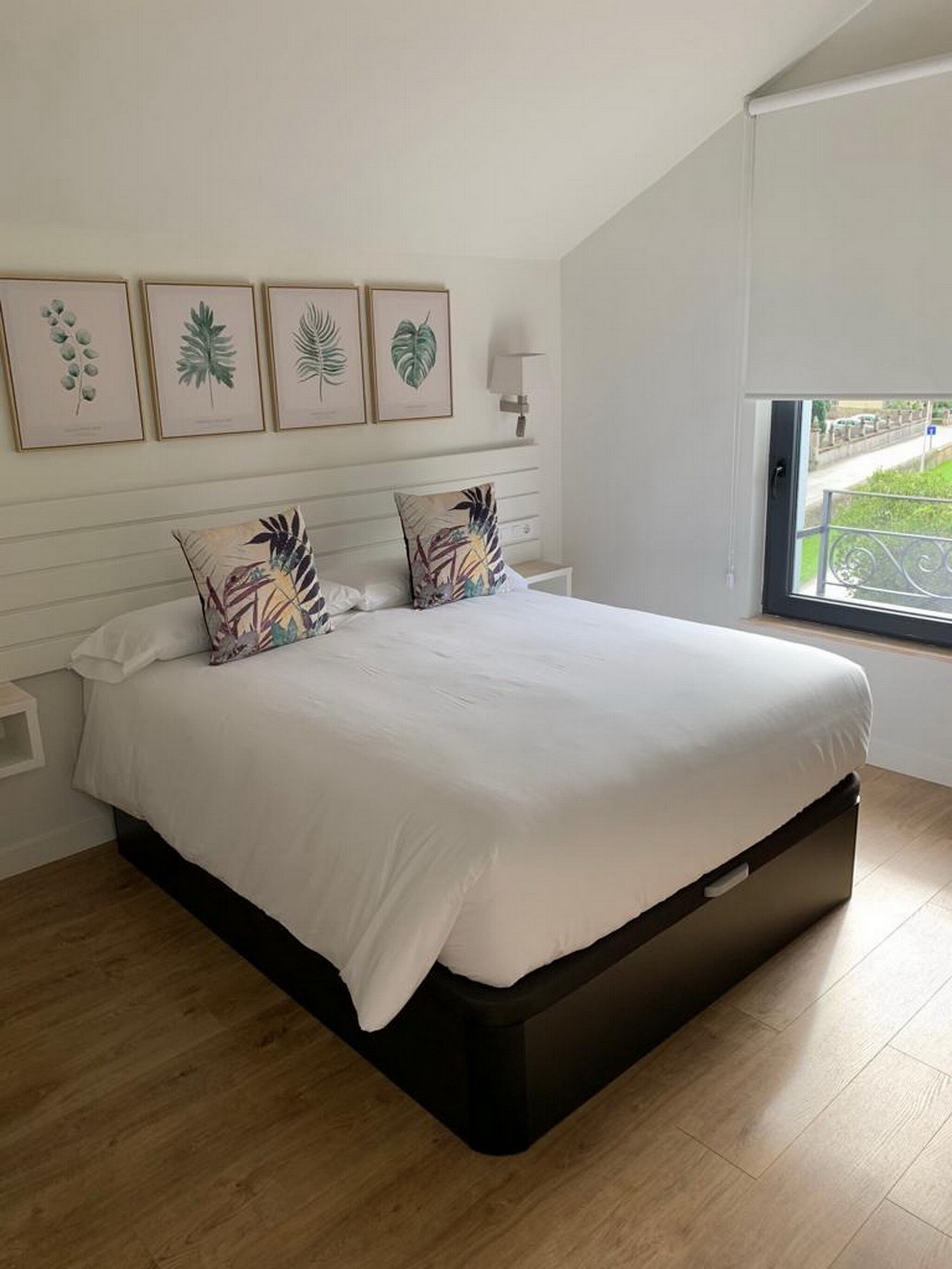 A modern bedroom with a large bed, wooden flooring, and a window with a view of greenery.