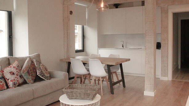 Een moderne woonkamer met een bank, stoelen, een eettafel en een keuken met witte kasten en een hanglamp.