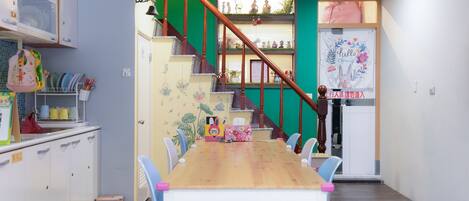 A dining area with a wooden table and blue chairs, a staircase with a wooden railing, and a wall with a 'Welcome' sign.