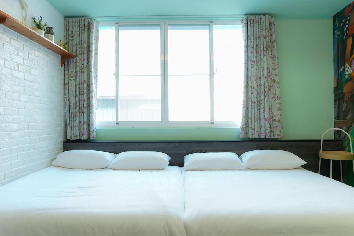 A room with two beds, a window with floral curtains, a brick wall, and a green wall.