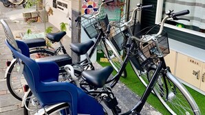 Eine Reihe von Stillfahrrädern mit Körben, ein blauer Stuhl und ein Roller vor einem Gebäude mit blauer Wand und einem Bereich mit künstlichem Rasen.