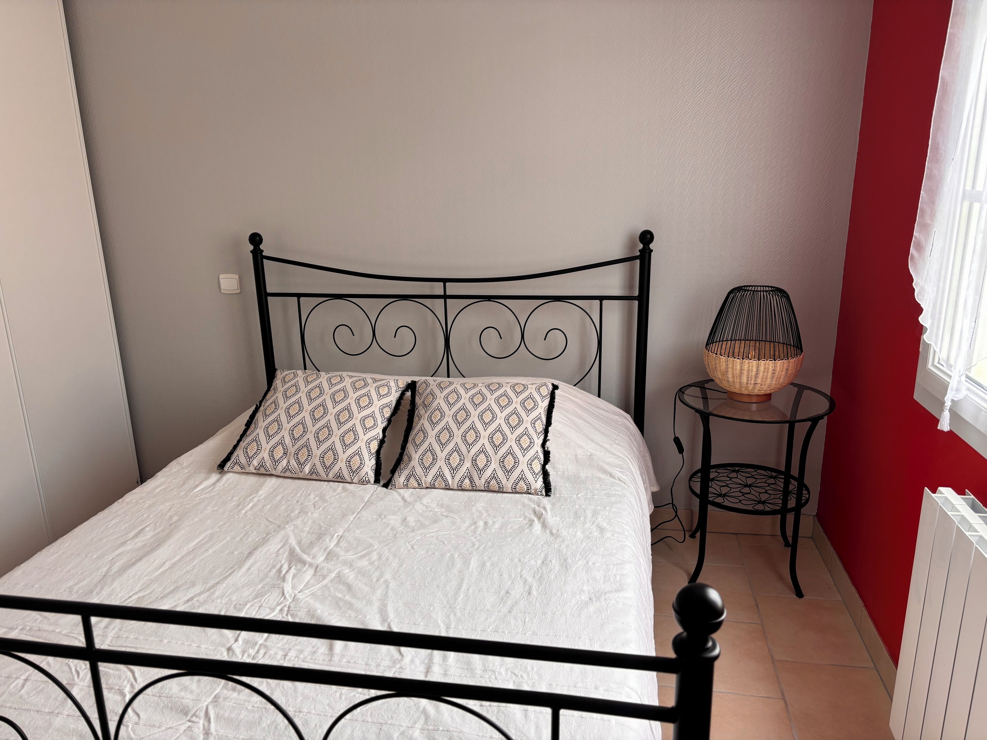 A bed with a metal frame, white bedding, and patterned pillows. There is a small round table with a wicker lamp beside the bed.