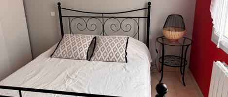 A bed with a metal frame, white bedding, and patterned pillows. There is a small round table with a wicker lamp beside the bed.