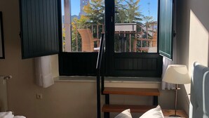 A bedroom with a bed, pillows, a nightstand, a lamp, and a window with open shutters. - Hotel La Muralla (Zafra)