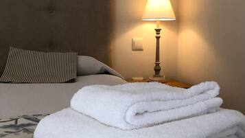 A neatly made bed with a floral-patterned bedspread, a folded white towel, a lamp on the nightstand, and a pillow with a striped pattern.