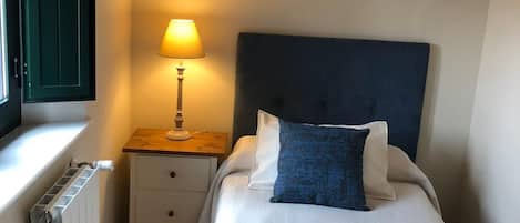 Un dormitorio ordenado con cama, mesita de noche con lámpara, radiador y ventana con persianas cerradas.