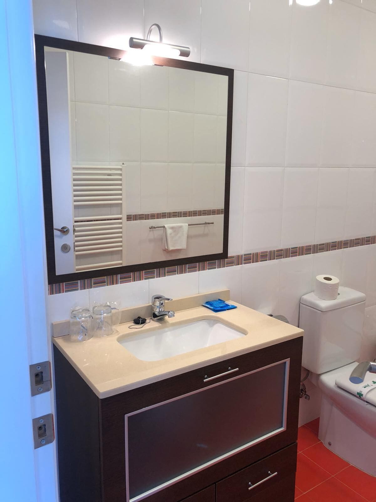 A bathroom with a sink, mirror, towel rack, toilet, and a small trash bin.
