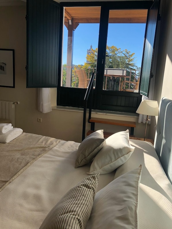A bedroom with a bed, pillows, a window with a view of trees and a building, a wooden bench, and a lamp. - Hotel La Muralla (Zafra)