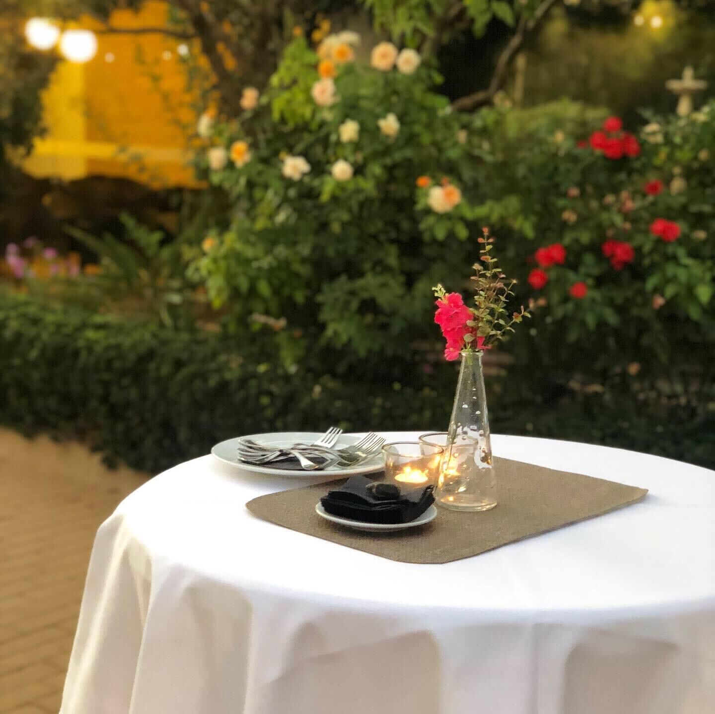 Une table ronde installée à l’extérieur, recouverte d’une nappe blanche, agrémentée d’un vase en verre transparent rempli de fleurs et d’une bougie.
