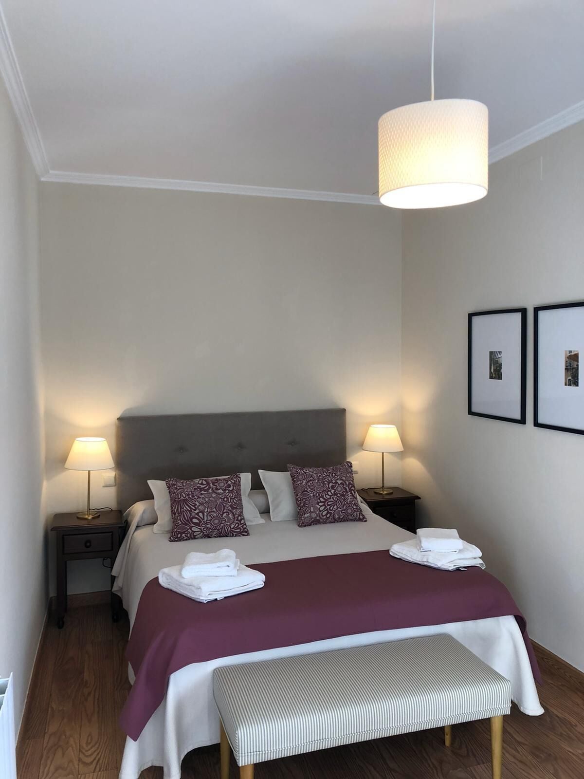 A neatly arranged hotel room with a bed, bedside lamps, a bench, and two framed pictures on the wall.