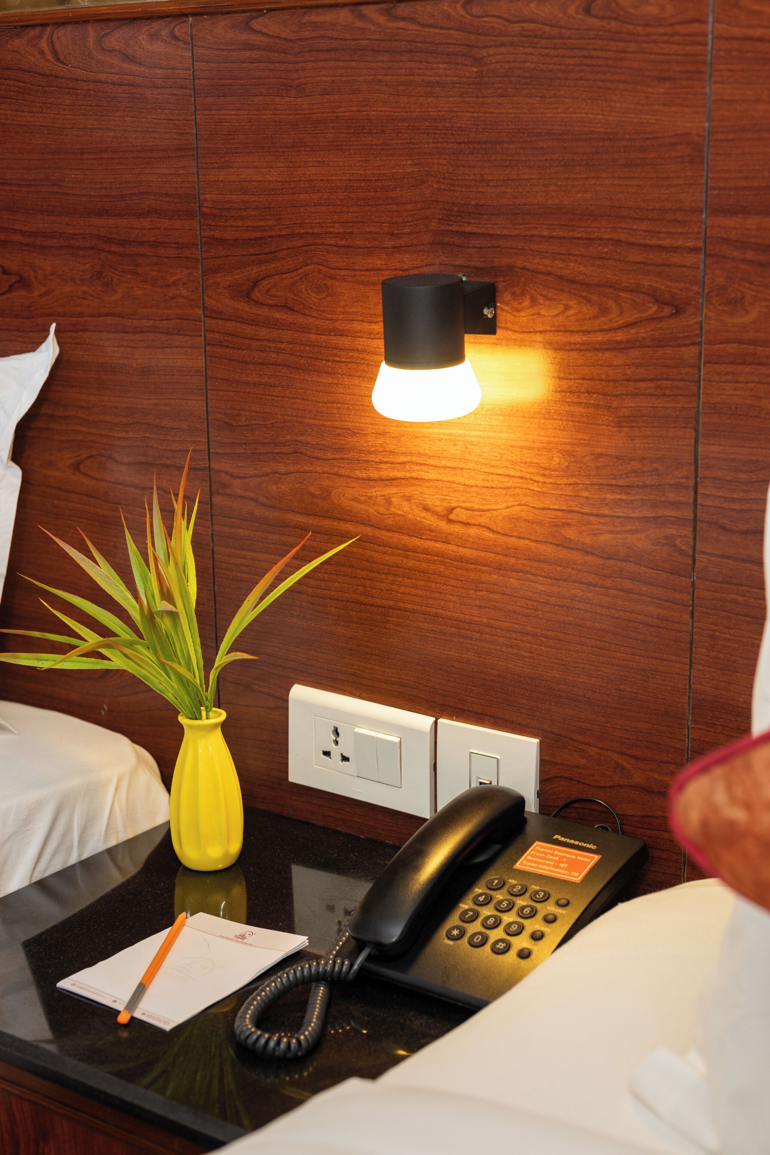 A hotel room with a wooden headboard, a bedside table with a yellow vase, a black telephone, and a notepad.
