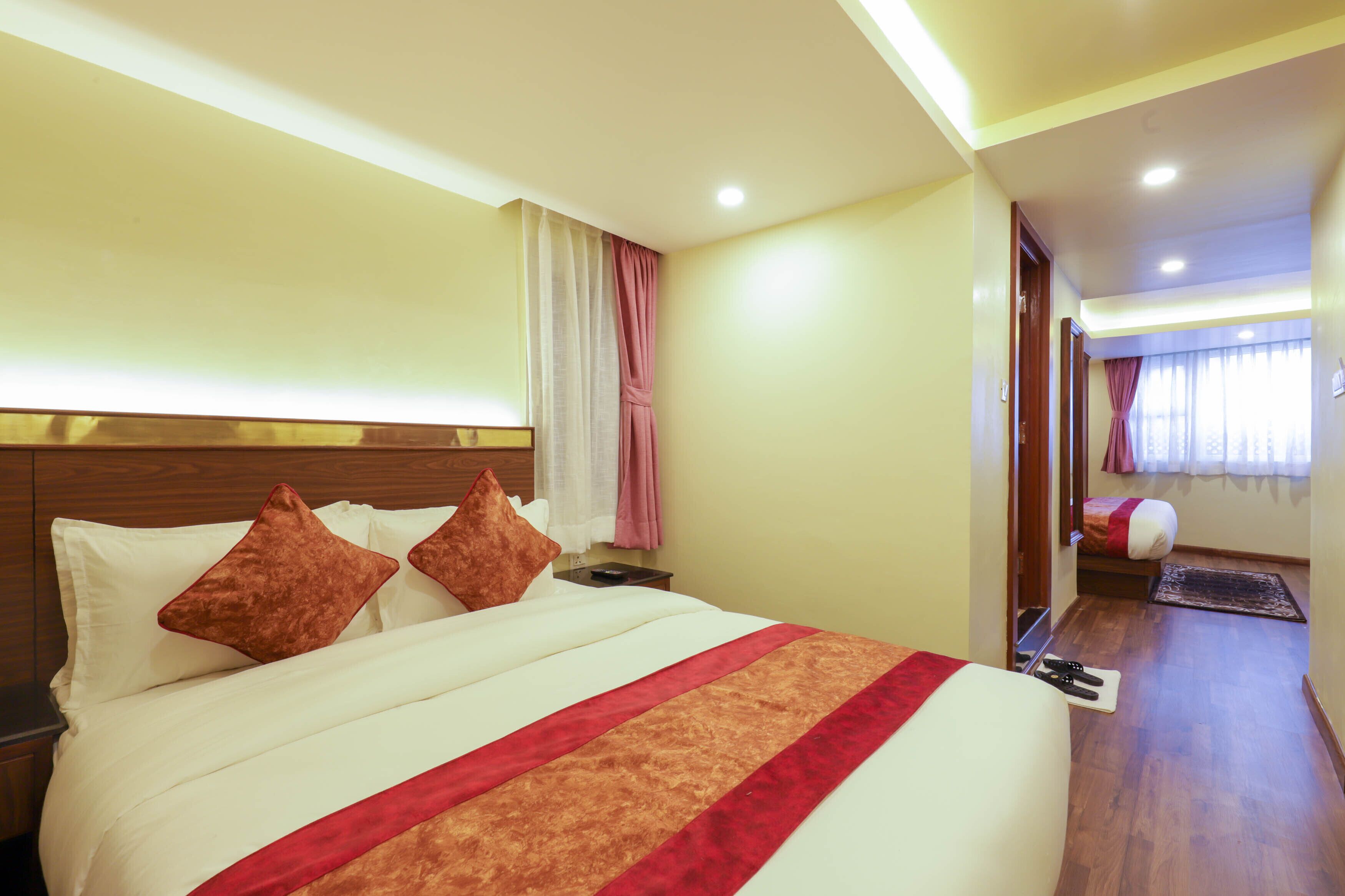 Habitación de hotel con una cama grande, una mesita de noche, un cabecero de madera, una ventana con cortinas y una puerta que da a otra habitación.