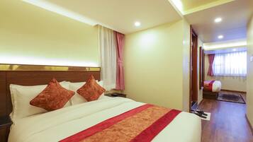 Habitación de hotel con una cama grande, una mesita de noche, un cabecero de madera, una ventana con cortinas y una puerta que da a otra habitación.