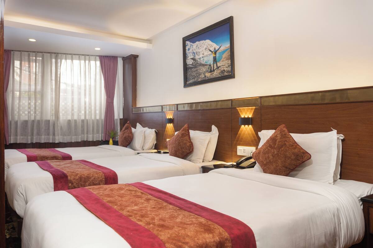 Ein Hotelzimmer mit zwei Betten, jeweils mit einer roten und braunen Tagesdecke, weißen Kissen und einem hölzernen Kopfteil. An der Wand hängt ein Gemälde einer Berglandschaft, und es gibt ein Fenster mit durchsichtigen Vorhängen.