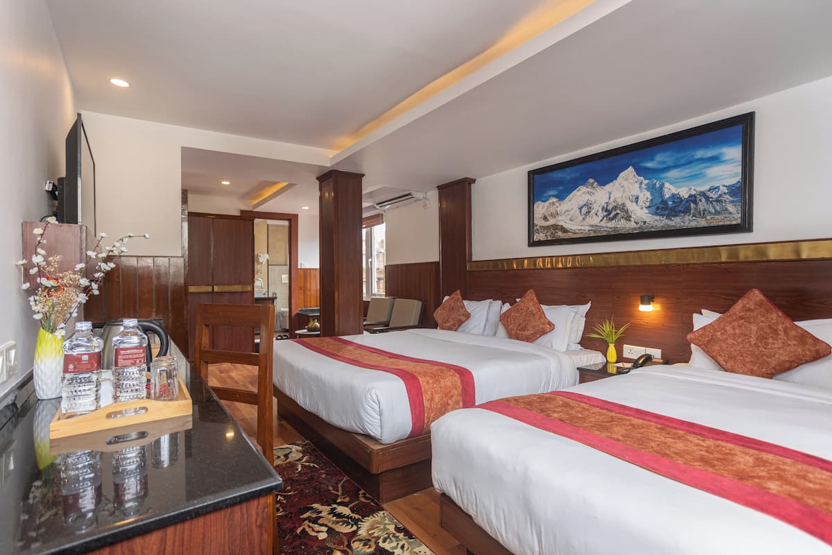 Ein Hotelzimmer mit zwei Betten, einem Schreibtisch mit Blumenvase und einem Bergpanorama an der Wand.