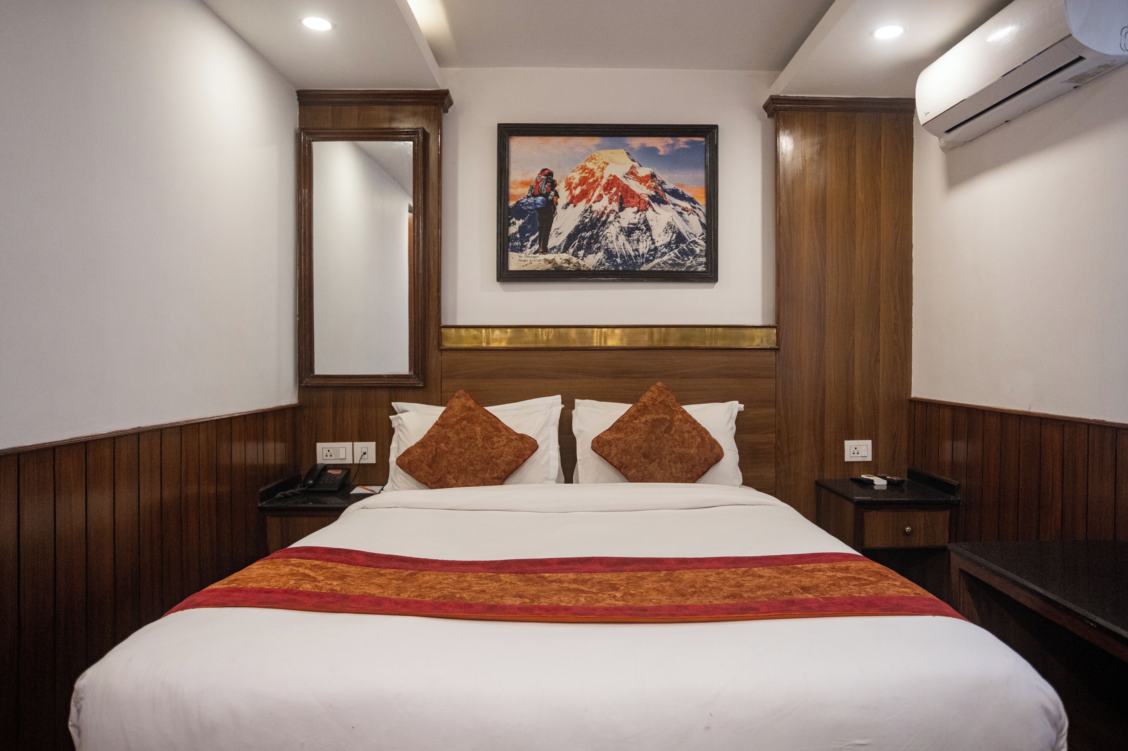 Habitación de hotel con una cama grande, paneles de madera, un cuadro de una montaña, una mesita de noche y un equipo de aire acondicionado.
