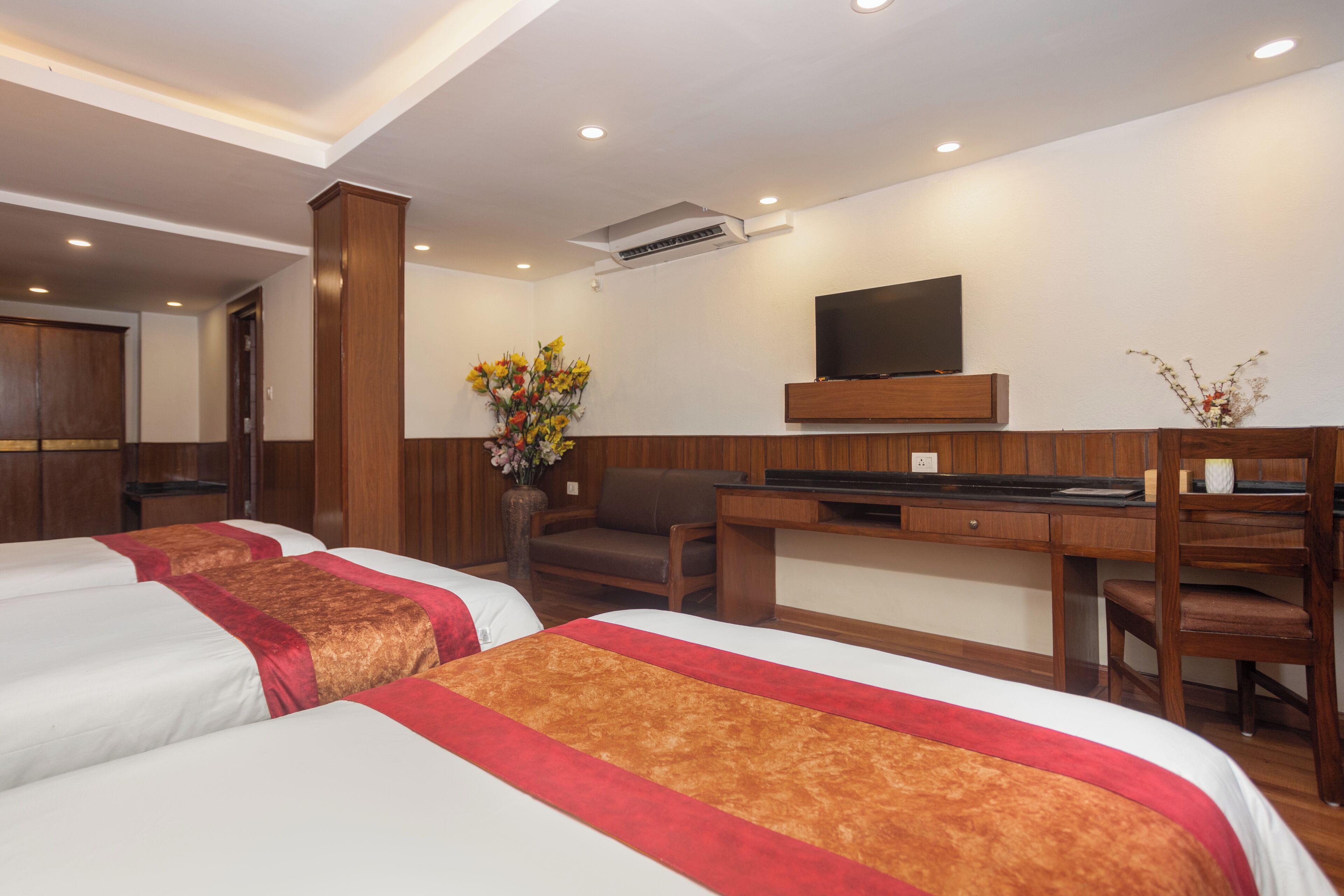 A hotel room with two beds, a desk, a chair, a TV, and a vase with flowers.