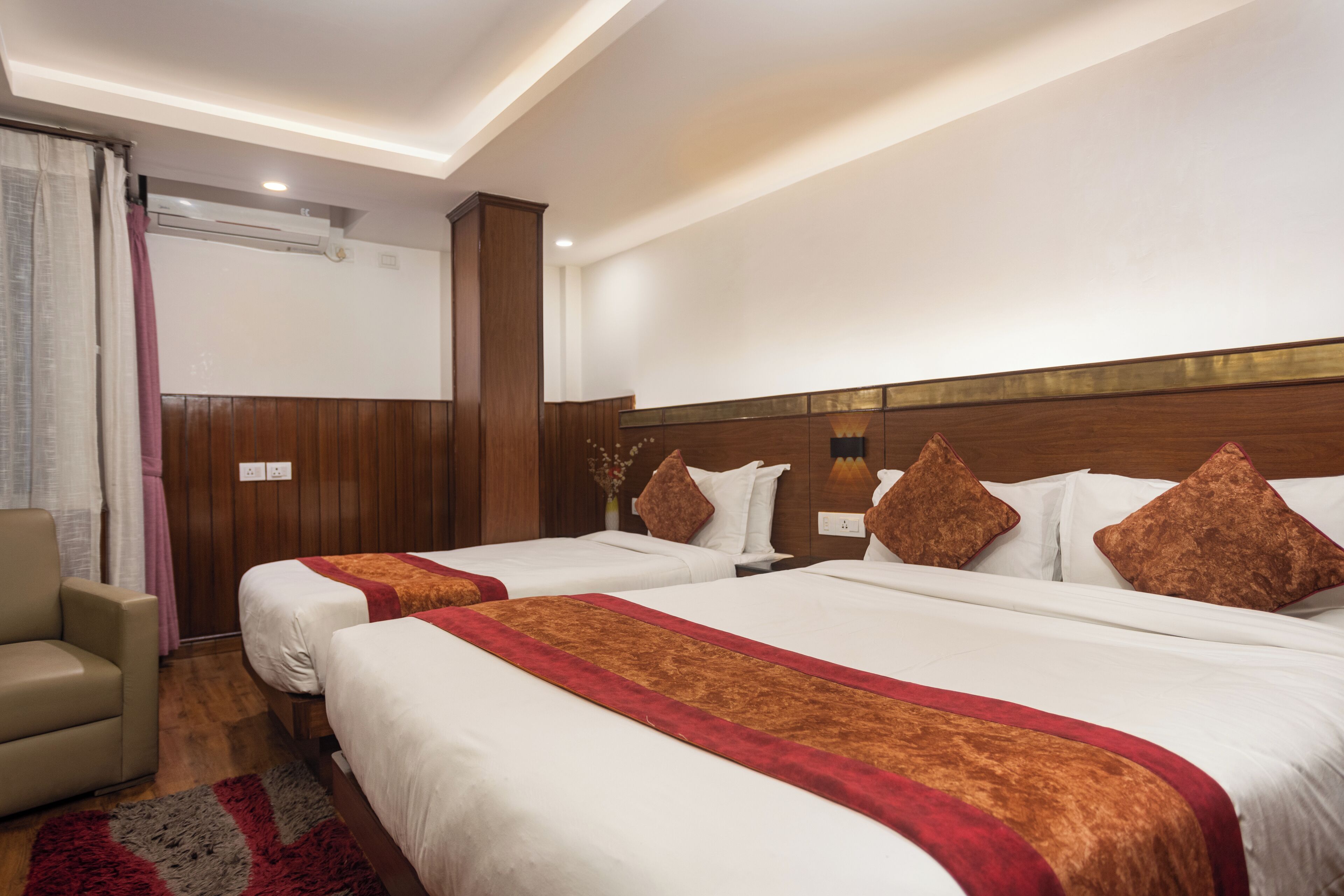 A hotel room with two beds, a wooden headboard, a red and white bedspread, a grey and red rug, and a beige armchair.