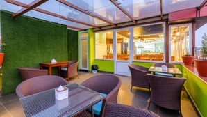 Uma área de estar coberta ao ar livre com móveis de vime e uma mesa de vidro, ao lado de um restaurante com parede verde e portas de vidro.