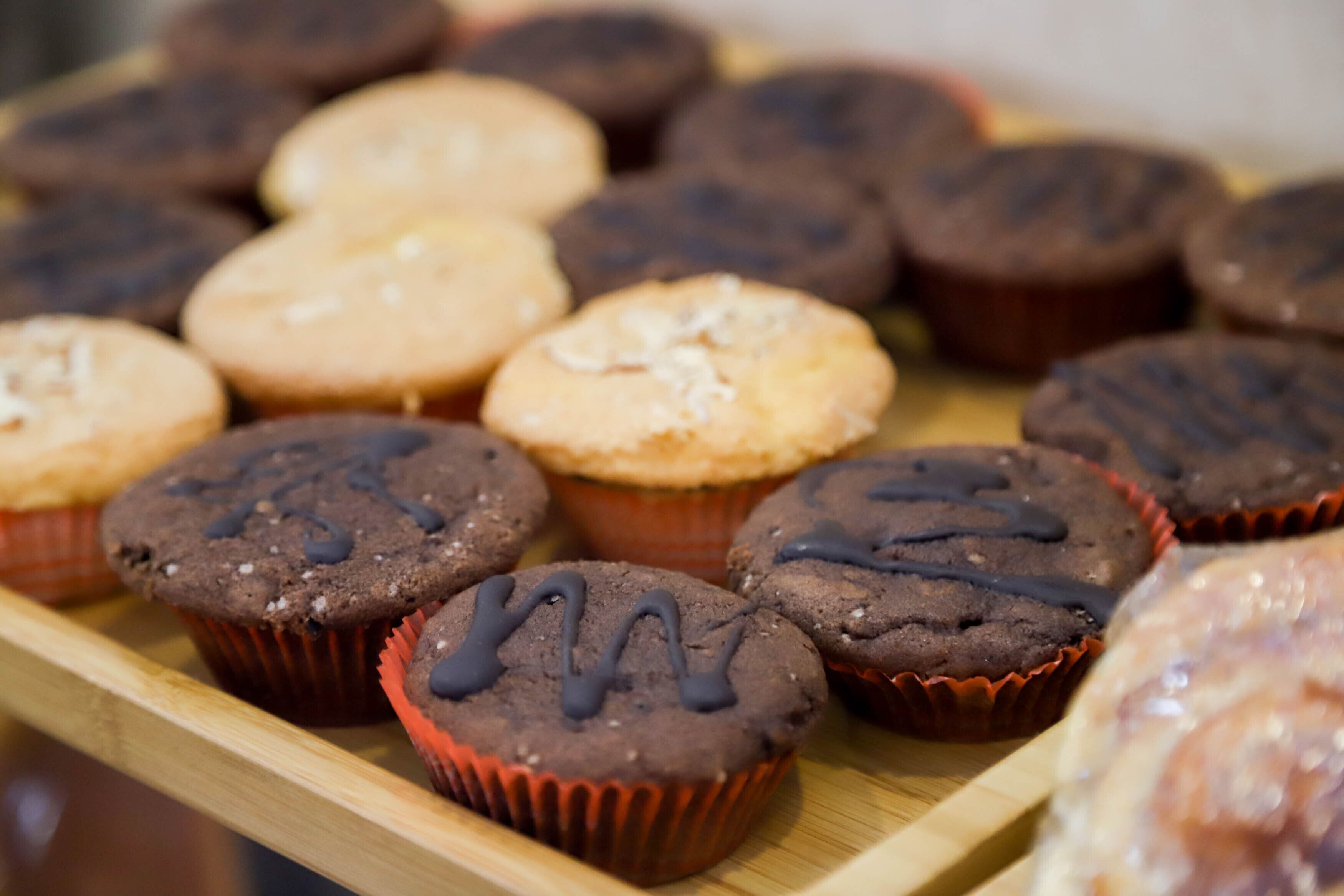 A wooden tray with various muffins, some with chocolate drizzle and others with a dusting of powdered sugar.