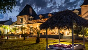 A thatched-roof outdoor seating area with a cushioned bench, surrounded by a well-maintained lawn and potted plants, in front of a building with multiple chimneys and a dark, cloudy sky. - Domaine St Ferreol (Chis)
