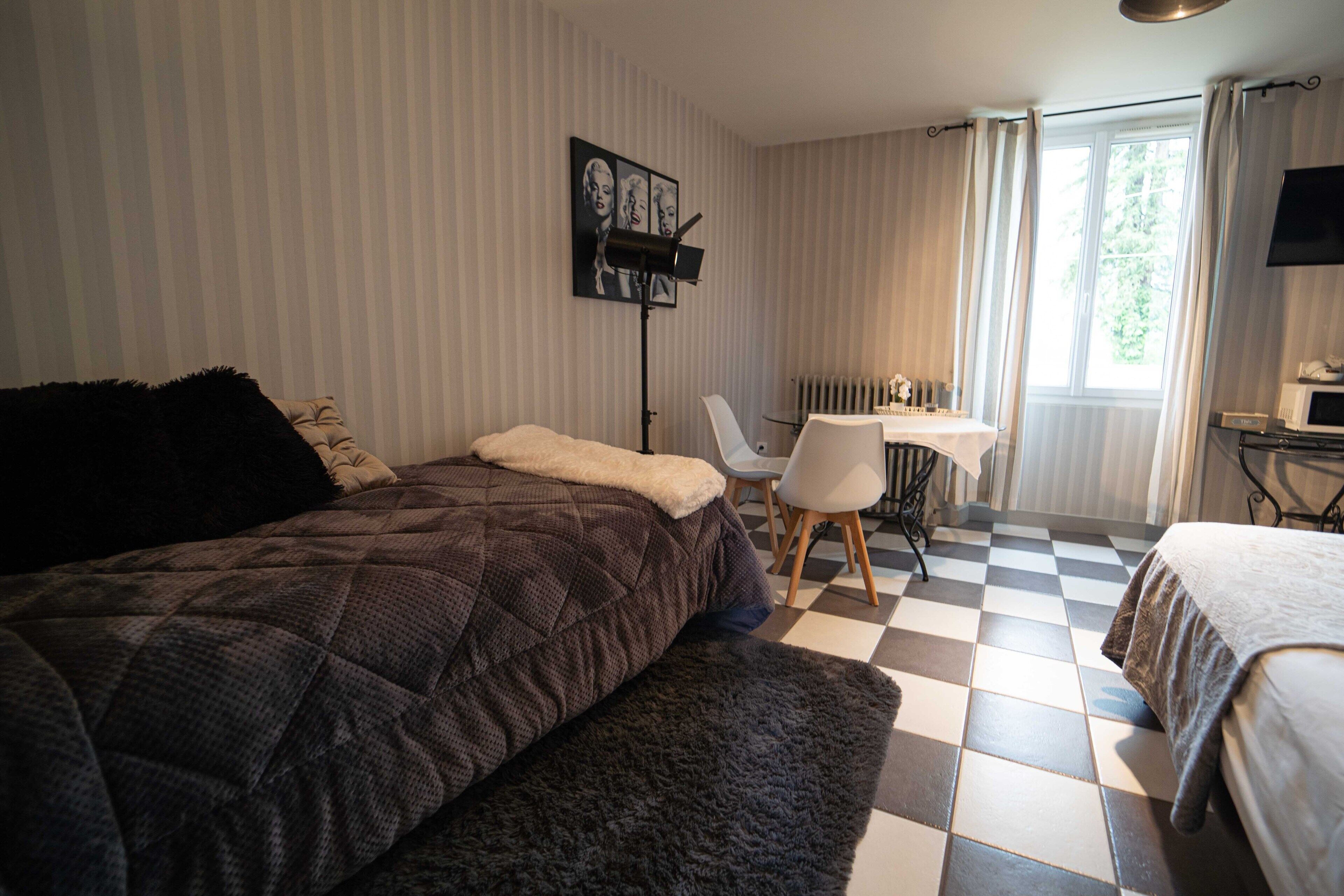 Ett hotellrum med ett rutigt golv, en säng med ett mörkt täcke, ett skrivbord med en stol och ett fönster med gardiner.