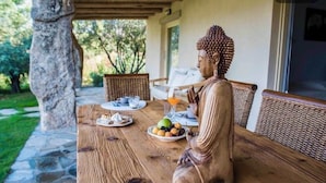 Een Boeddhabeeld zittend aan een houten tafel met een schaal fruit en een glas sinaasappelsap.