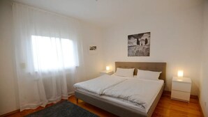 A bedroom with a bed, two bedside tables, a nightstand, a window with sheer curtains, a framed picture on the wall, and a dark-colored rug. - WeinheimResidence (2) (Weinheim)