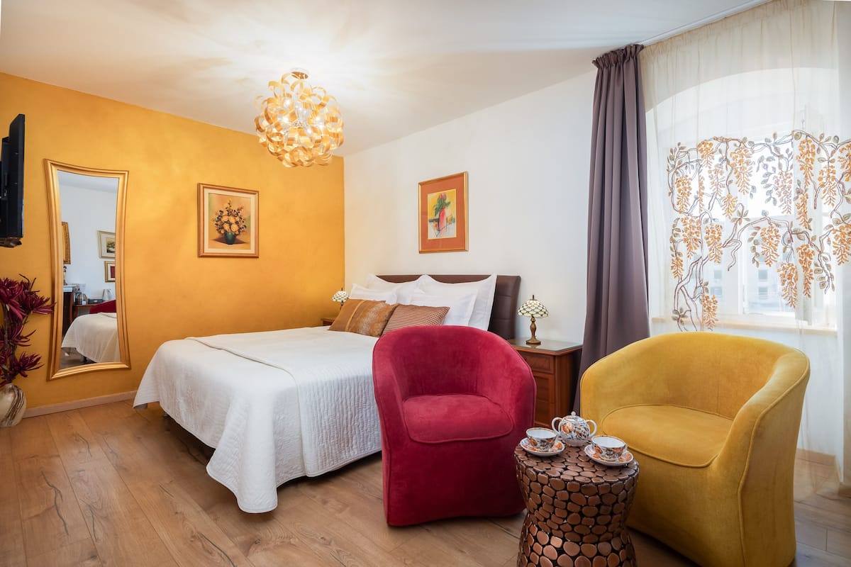 A bedroom with a bed, a red chair, a yellow armchair, a small table with a teapot, and a mirror on the wall.