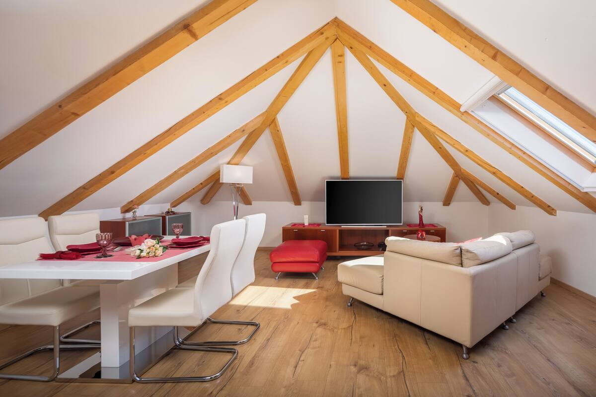 A loft-style room with a dining table, sofa, TV, and wooden beams.