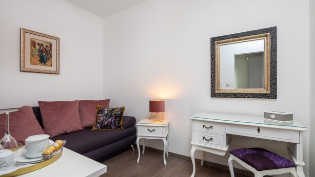 A modern living room with a sofa, a glass coffee table with a tray of items, a white side table with a lamp, a white desk with drawers, and a large mirror on the wall.