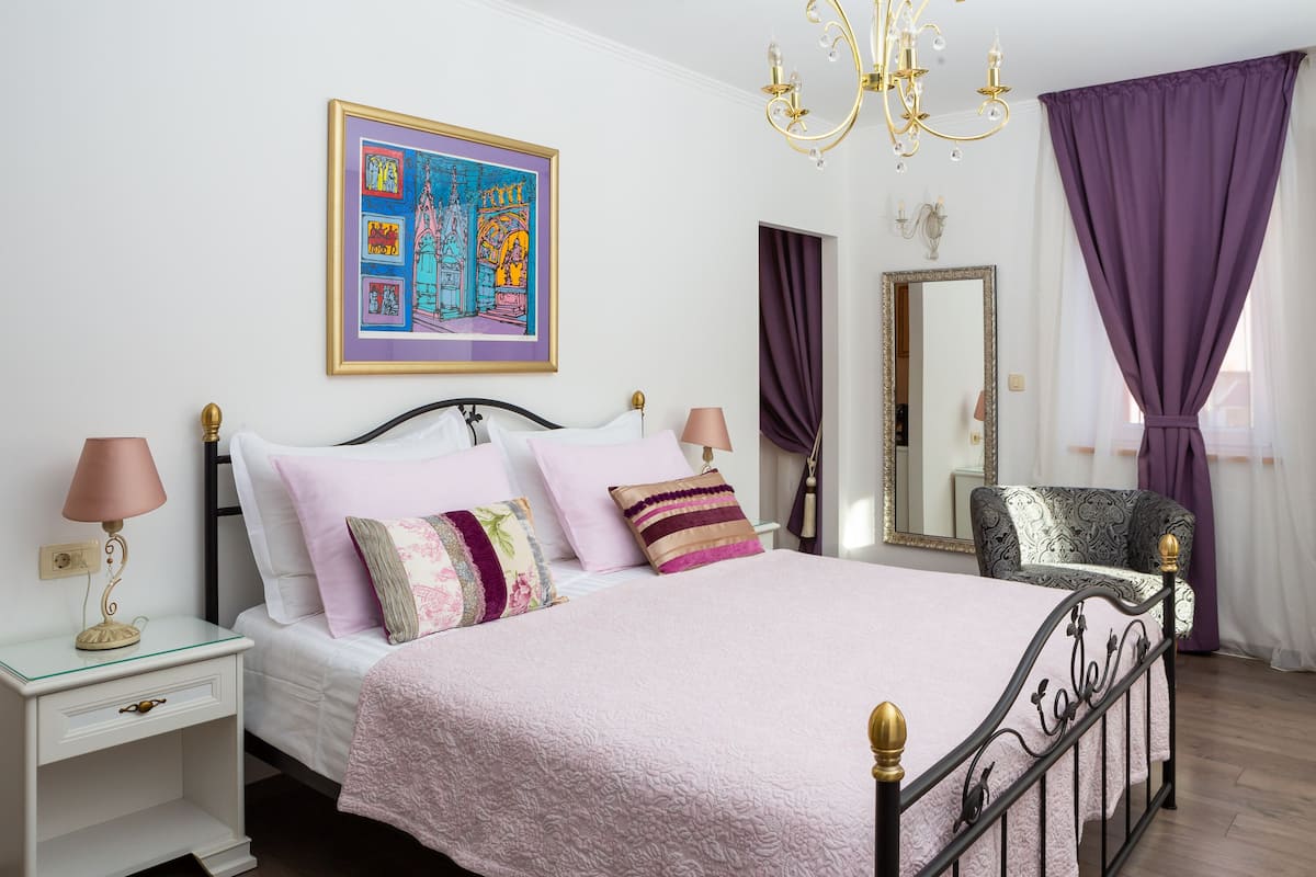 A neatly arranged bedroom with a bed, bedside tables, a chair, a mirror, and a chandelier.