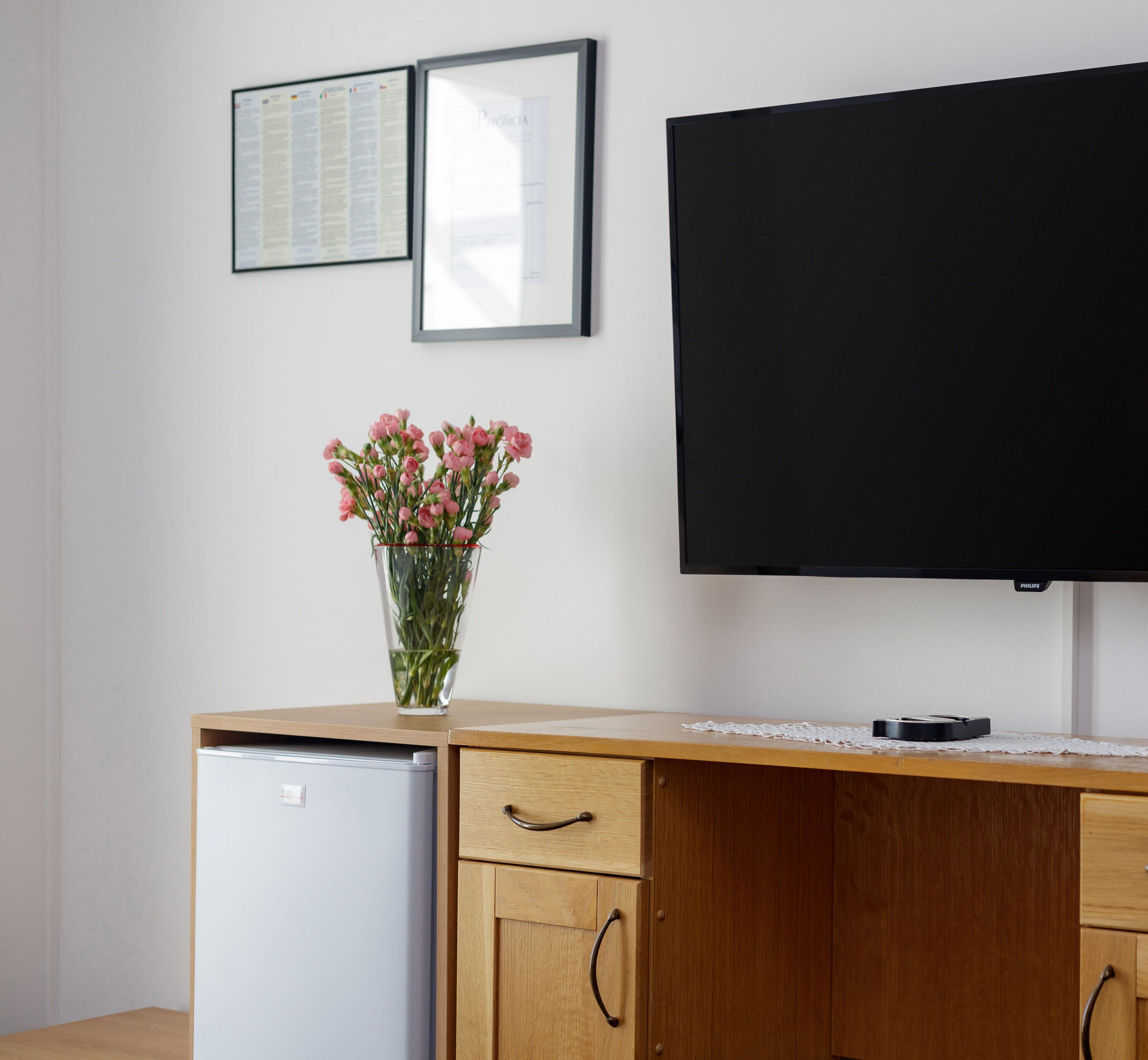 A kitchenette with a refrigerator, a wooden cabinet with a drawer, a vase with flowers, and a flat-screen TV mounted on the wall.