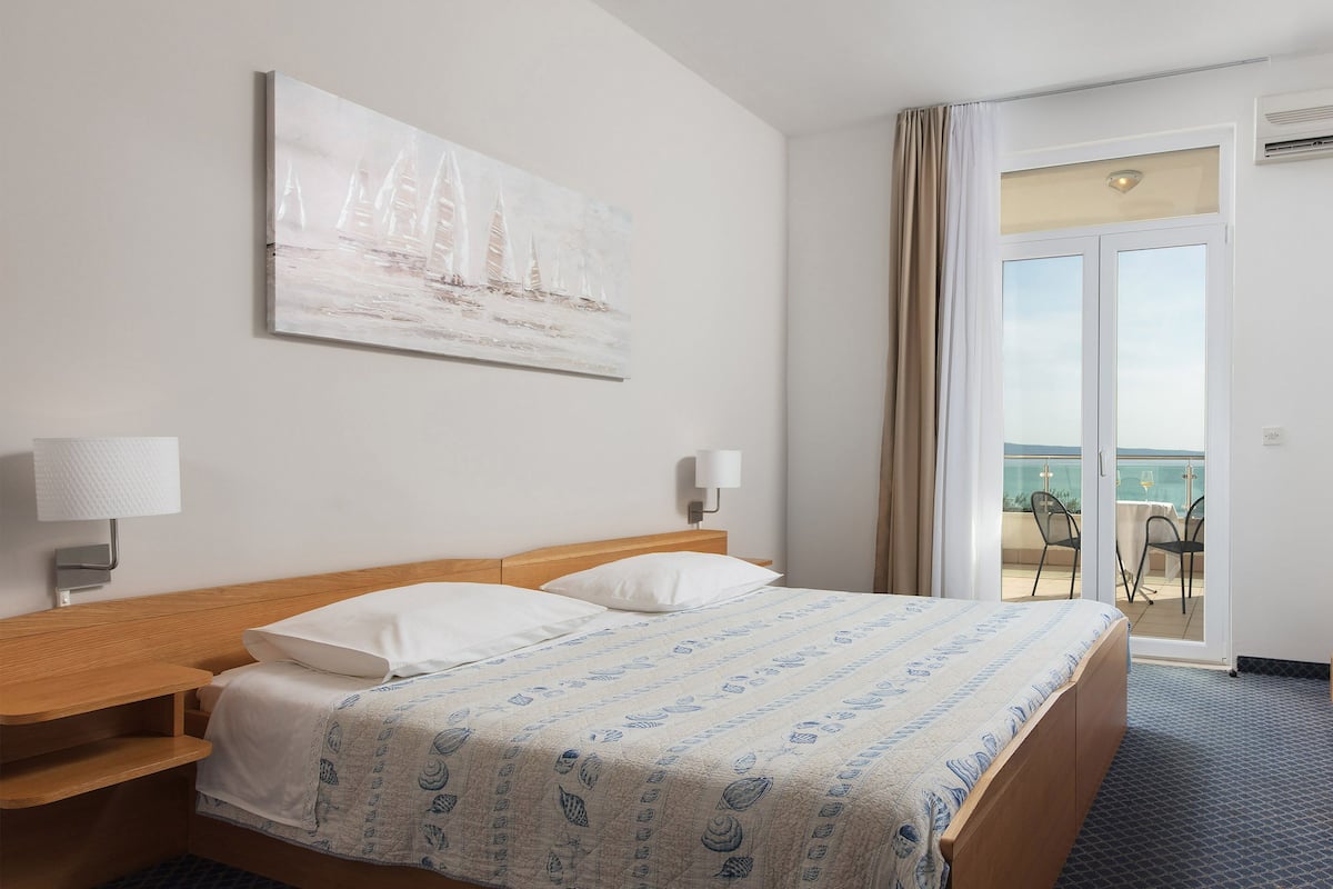 A hotel room with a bed, bedside tables, a painting, and a view of the sea through a sliding door.