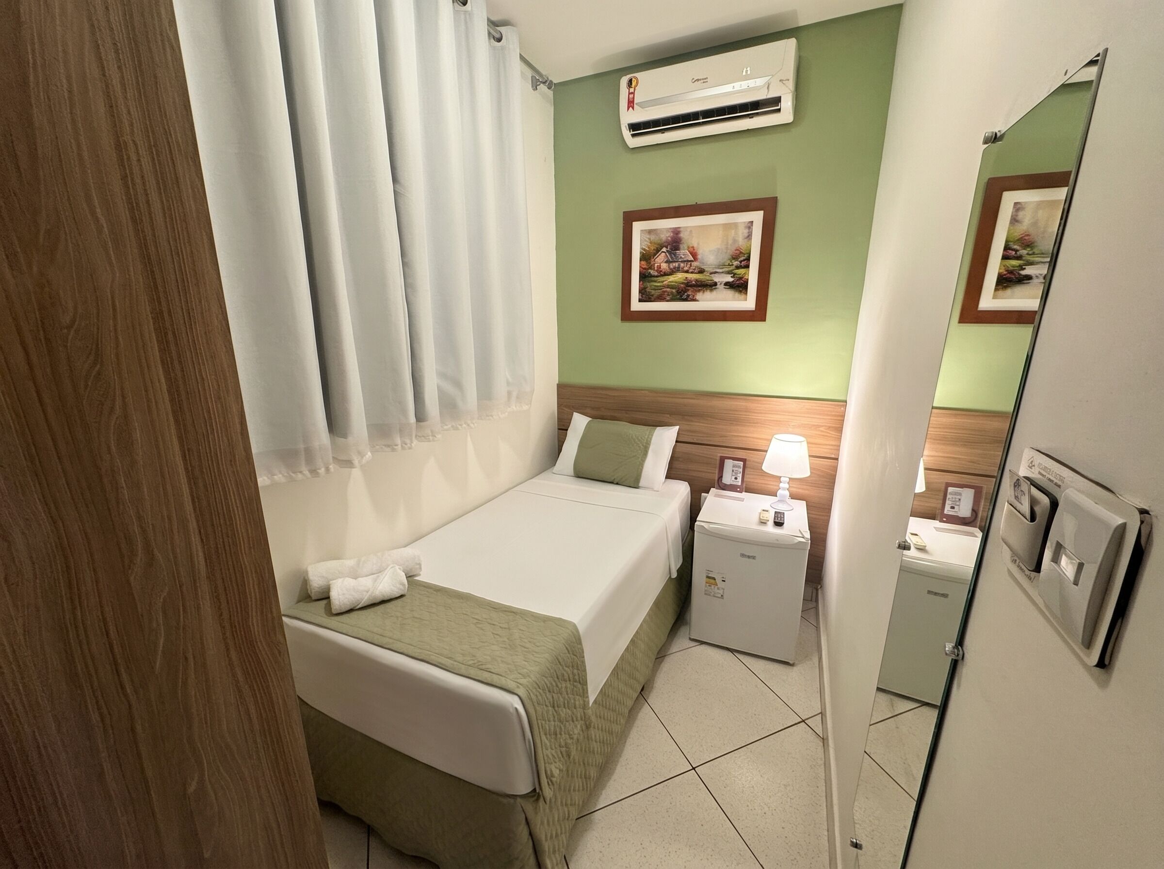 A hotel room with a bed, bedside table, lamp, air conditioning unit, and a mirror.