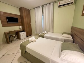 Ein Hotelzimmer mit zwei Betten, einem Schreibtisch mit Stuhl, einem Fernseher, einer Klimaanlage und einem Fenster mit Vorhängen.