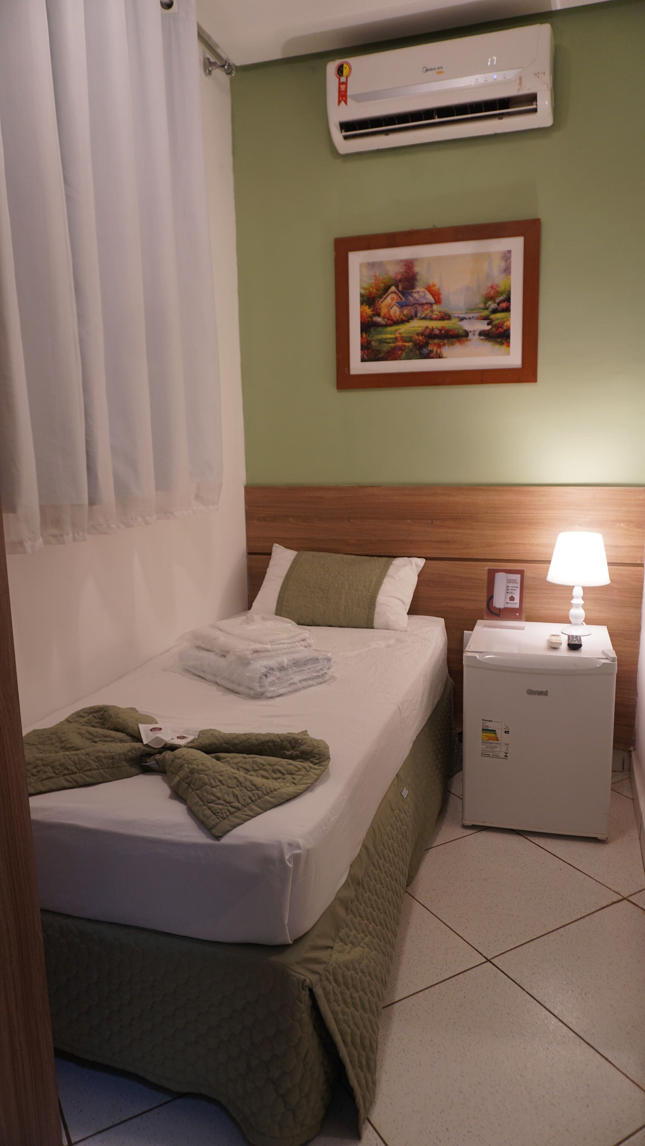 A hotel room with a bed, a nightstand, a lamp, an air conditioner, and a painting on the wall.