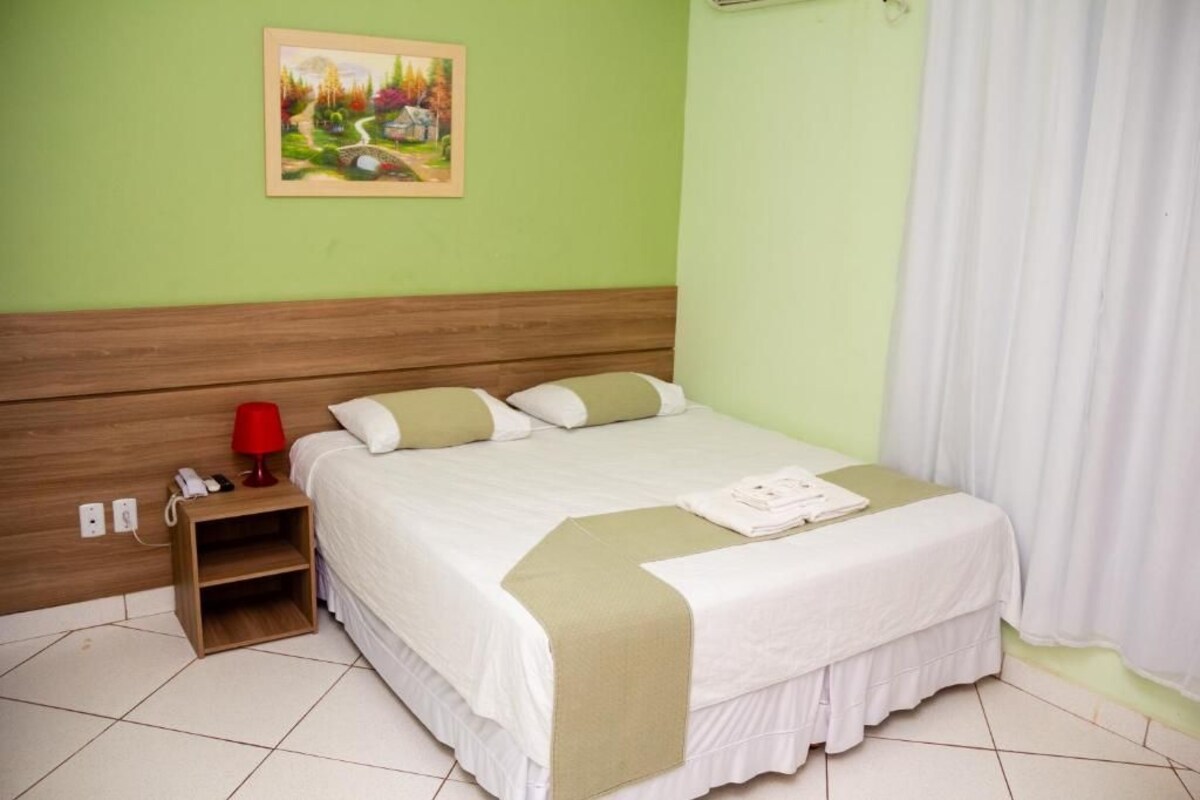 Una cama bien tendida con colcha blanca y verde, una lámpara roja y una mesita de noche de madera con un teléfono y una lámpara.
