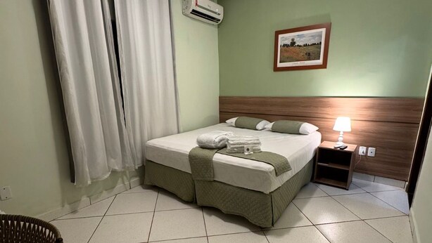Een opgemaakt bed met een groen hoofdeinde, een nachtkastje met een lamp, en een ingelijst foto op de muur.
