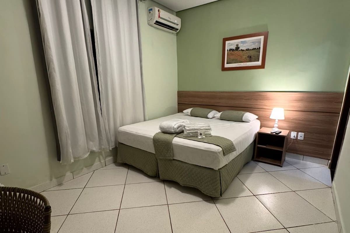 Un lit bien fait, avec une tête de lit verte, une table de chevet sur laquelle se trouve une lampe, et un tableau encadré accroché au mur.