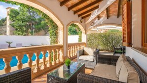 A covered outdoor area with wicker furniture, a glass table, and a view of a pool and greenery. - Villa Can Odile By SunVillas Mallorca (Cala San Vicens)