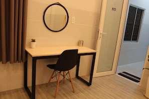 A small, modern room with a desk, a chair, a round mirror, a potted plant, and a door with a window. - B Home - Hostel (Can Tho)