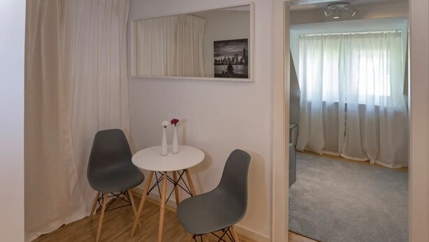 Une pièce avec un plancher en bois, deux chaises grises, une petite table blanche et un vase blanc rempli de fleurs. Un tableau encadré est accroché au mur, et une porte donne accès à une autre pièce.