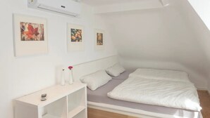 Une petite chambre moderne avec un lit de style industriel, des murs blancs et un plancher en bois. On y trouve trois tableaux encadrés, une petite console blanche et une bibliothèque.